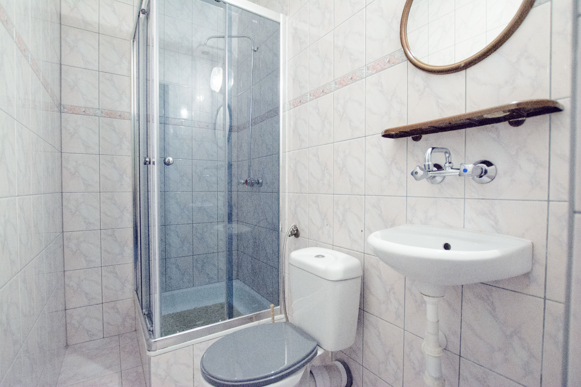 Jasna łazienka z kabiną prysznicową, toaletą z szarą deską, umywalką i okrągłym lustrem z drewnianą półką.