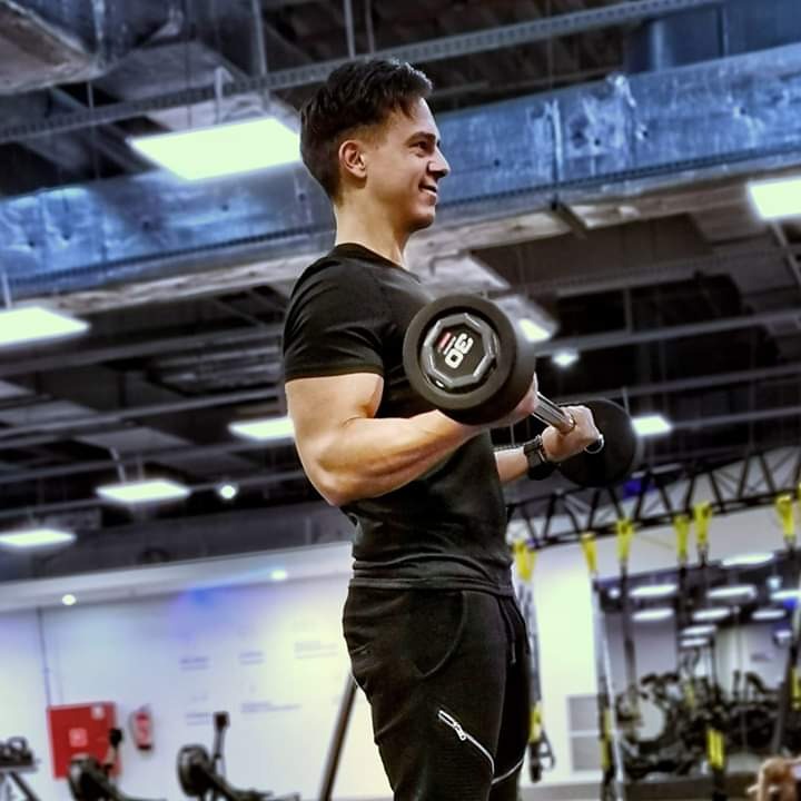 Uśmiechnięty mężczyzna w czarnym stroju ćwiczy biceps z obciążeniem 30 kg w siłowni z surowym, industrialnym sufitem.