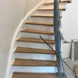 Schody wewnętrzne z drewnianymi stopniami i pomalowanymi na biało podstopnicami, szara metalowa balustrada, widoczne ślady prac wykończeniowych na ścianie i podłodze.