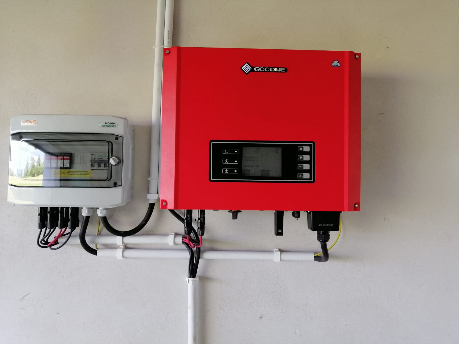 Czerwony inwerter GoodWe zamontowany na ścianie obok skrzynki bezpiecznikowej z widocznymi kablami i przewodami w białych rurkach PCV.