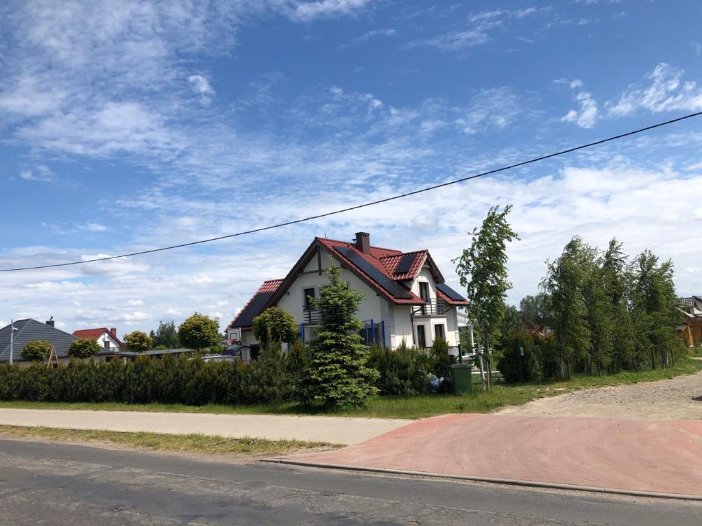 Dom jednorodzinny z czerwonym dachem i panelami na dachu, widok z ulicy w słoneczny dzień.