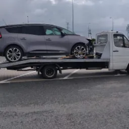 Uszkodzony szary samochód osobowy na platformie białej ciężarówki pomocy drogowej, zabezpieczony pasami transportowymi, na parkingu w pochmurny dzień.