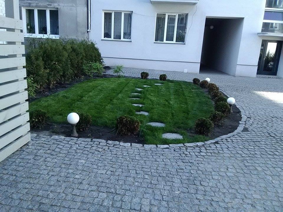 Ogródek z trawnikiem, ścieżką z kamieni, kulistymi lampami i obrzeżem z krzewów, otoczony kostką brukową przed nowoczesnym budynkiem.
