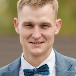 Portret młodego mężczyzny w jasnoniebieskiej marynarce i ciemnoniebieskiej muszce w kropki, z przypinką kwiatową, uśmiechającego się do kamery.