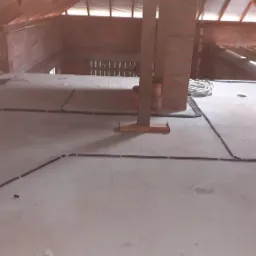 Surowy stan poddasza z widoczną instalacją elektryczną: czarne kable prowadzone po betonowej podłodze, przymocowane białymi uchwytami, zwój białych kabli w tle przy ceglanym kominie, widoczne...