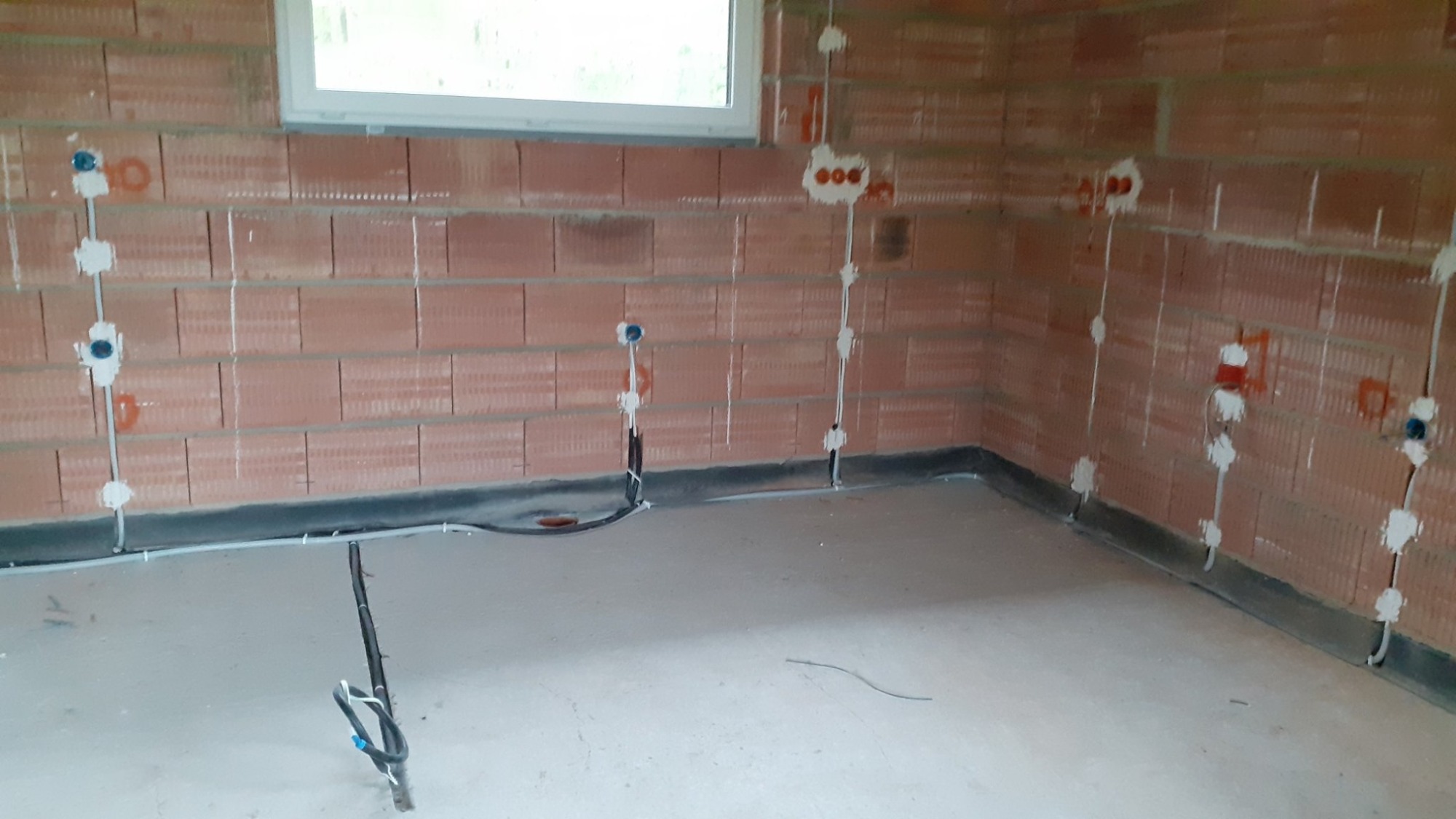 Surowy mur z instalacją elektryczną w trakcie montażu, widoczne kable i puszki pod gniazdka, okno w tle i surowa podłoga.