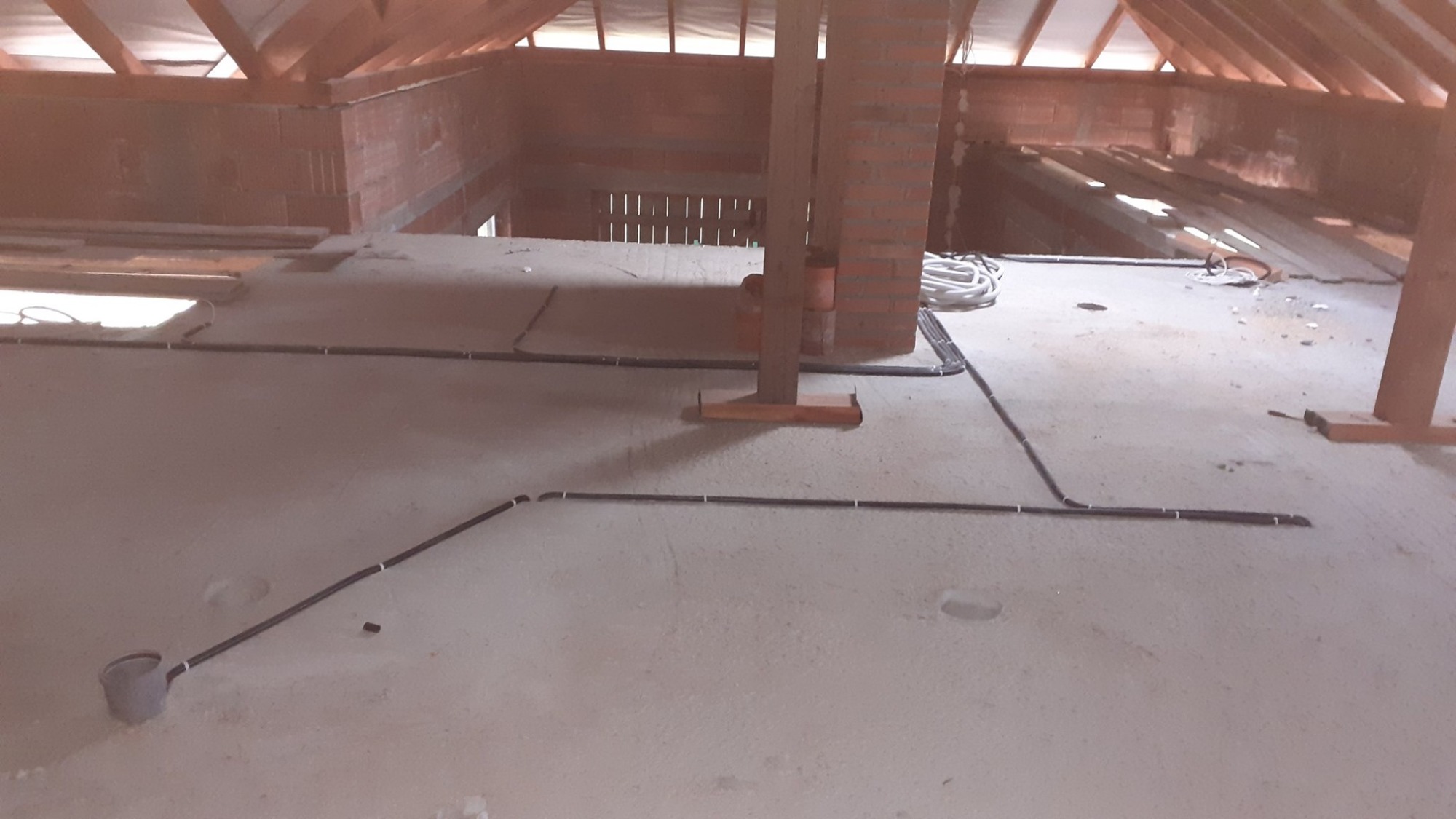 Surowy stan poddasza z widoczną instalacją elektryczną: czarne kable prowadzone po betonowej podłodze, przymocowane białymi uchwytami, zwój białych kabli w tle przy ceglanym kominie, widoczne...