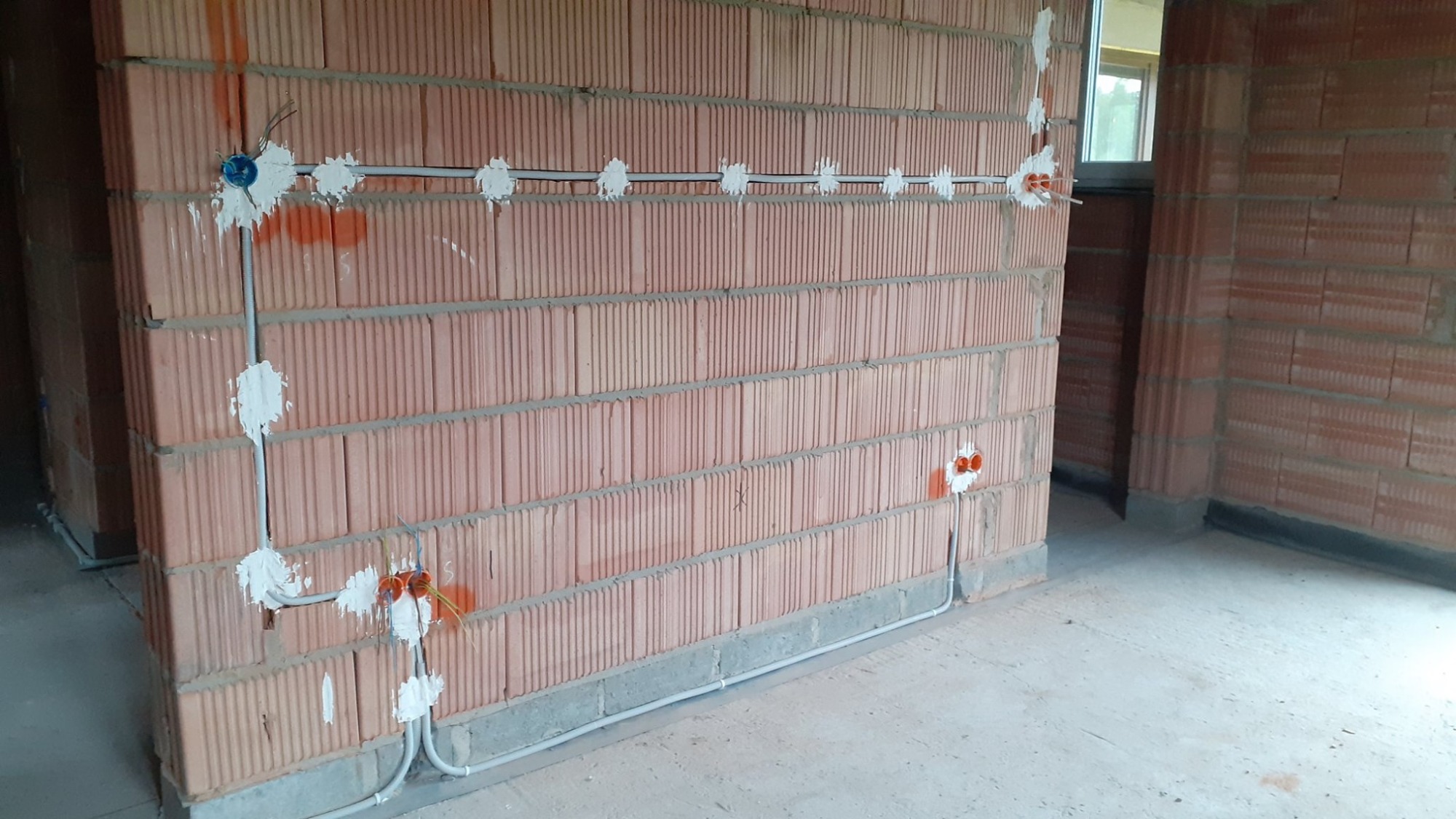 Surowa ceglana ściana z widocznymi przewodami elektrycznymi w szarych rurkach, zamocowanymi zaprawą gipsową, puszki instalacyjne w kolorze pomarańczowym i niebieskim, widoczne okablowanie.