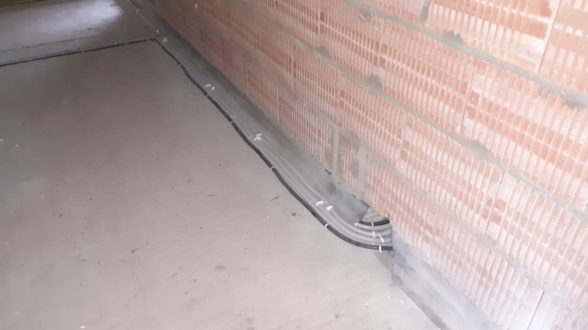 Szereg szarych rur instalacji elektrycznej przymocowanych do ceglanej ściany klamrami, widoczne na surowym betonowym podłożu.