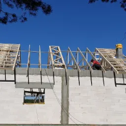 Budowa dachu na domu z białych bloczków, widoczny dekarz w czerwonej koszulce, drewniana konstrukcja dachu w trakcie montażu, komin z żółtym zabezpieczeniem.