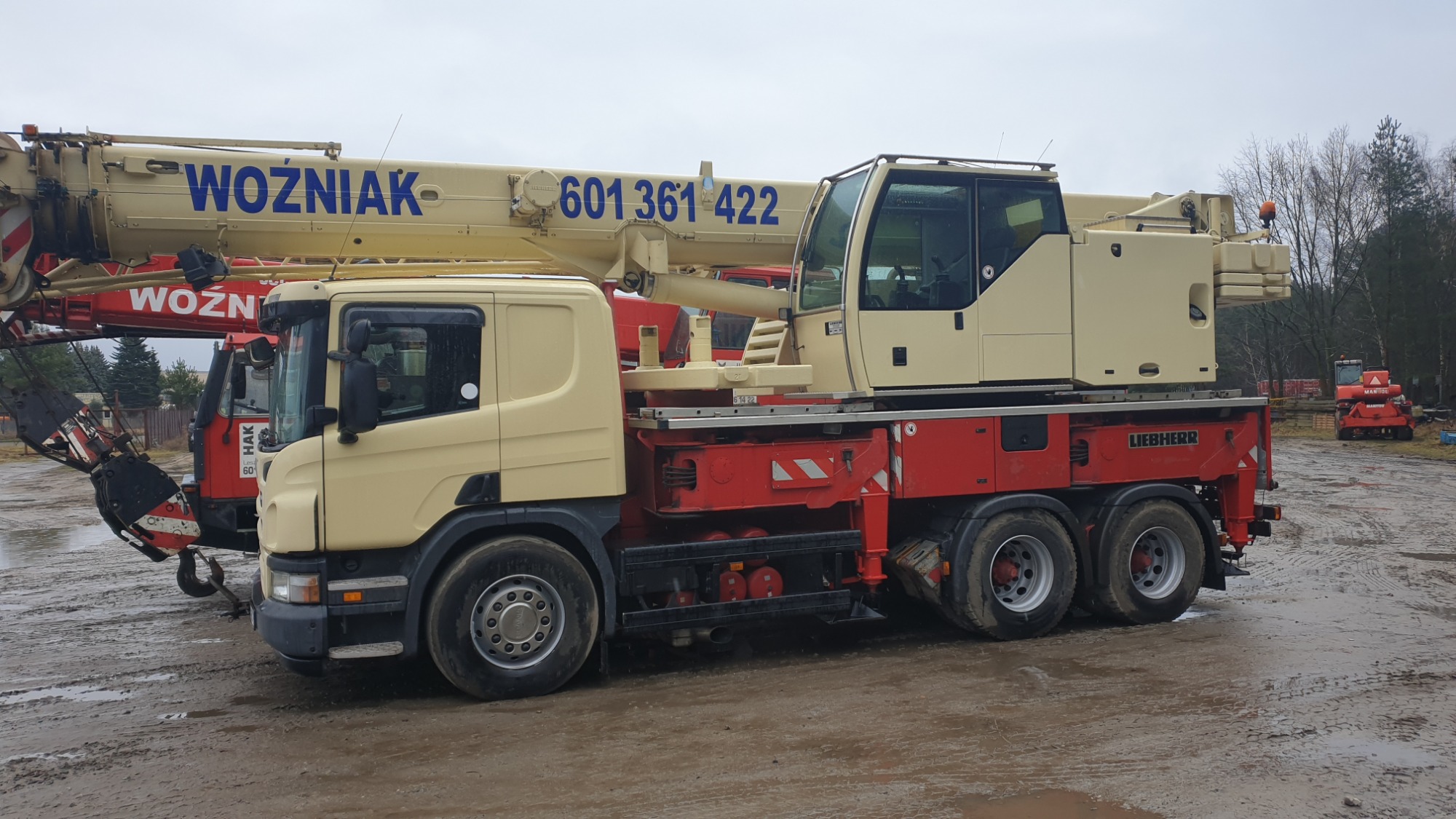 Żuraw samochodowy Liebherr w kolorach beżowo-czerwonych z napisem WOŹNIAK i numerem telefonu na wysięgniku, stojący na błotnistym terenie budowy, z widocznymi innymi maszynami budowlanymi w tle.