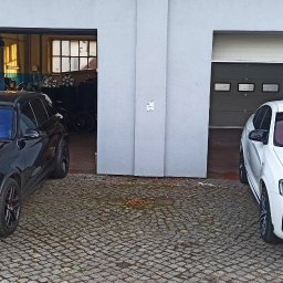 EXTREME DESIGN - Dwa samochody: czarny Mercedes-AMG GLC i białe BMW X4, zaparkowane przed garażami. Na szybach widoczne odbicia, sugerujące potencjalne przyciemnienie.