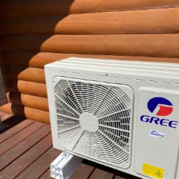 Zewnętrzna jednostka klimatyzacji marki Gree, zamontowana na drewnianym tarasie przy ścianie z bali, widoczna tabliczka ostrzegawcza i logo Inverter.