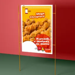 Plakat reklamowy na metalowym stojaku prezentujący smażonego kurczaka z hasłem 'Mega Chrupiący' i informacją o dostawie do domu, z ikonami telefonu i samochodu dostawczego.