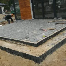 Budowa tarasu z szarej kostki brukowej na dwóch poziomach, widoczne narzędzia i pracownik w pomarańczowej czapce, częściowo ułożona nawierzchnia, piasek i obrzeża.