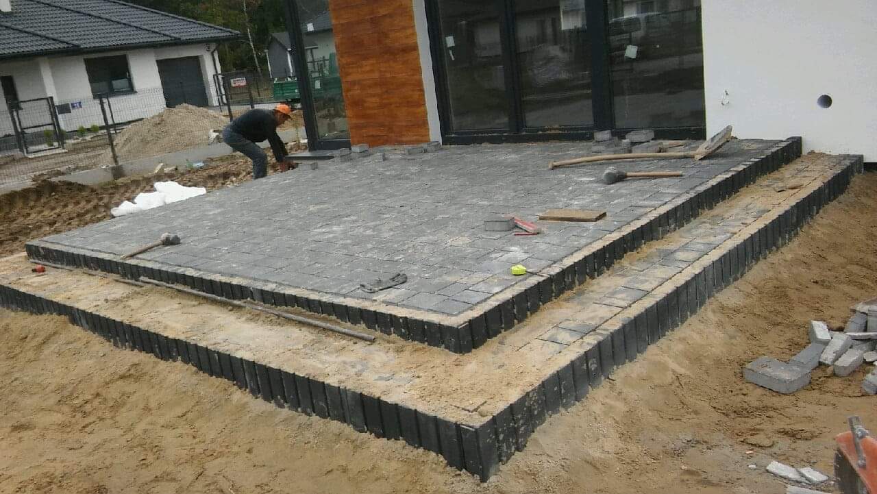 Budowa tarasu z szarej kostki brukowej na dwóch poziomach, widoczne narzędzia i pracownik w pomarańczowej czapce, częściowo ułożona nawierzchnia, piasek i obrzeża.