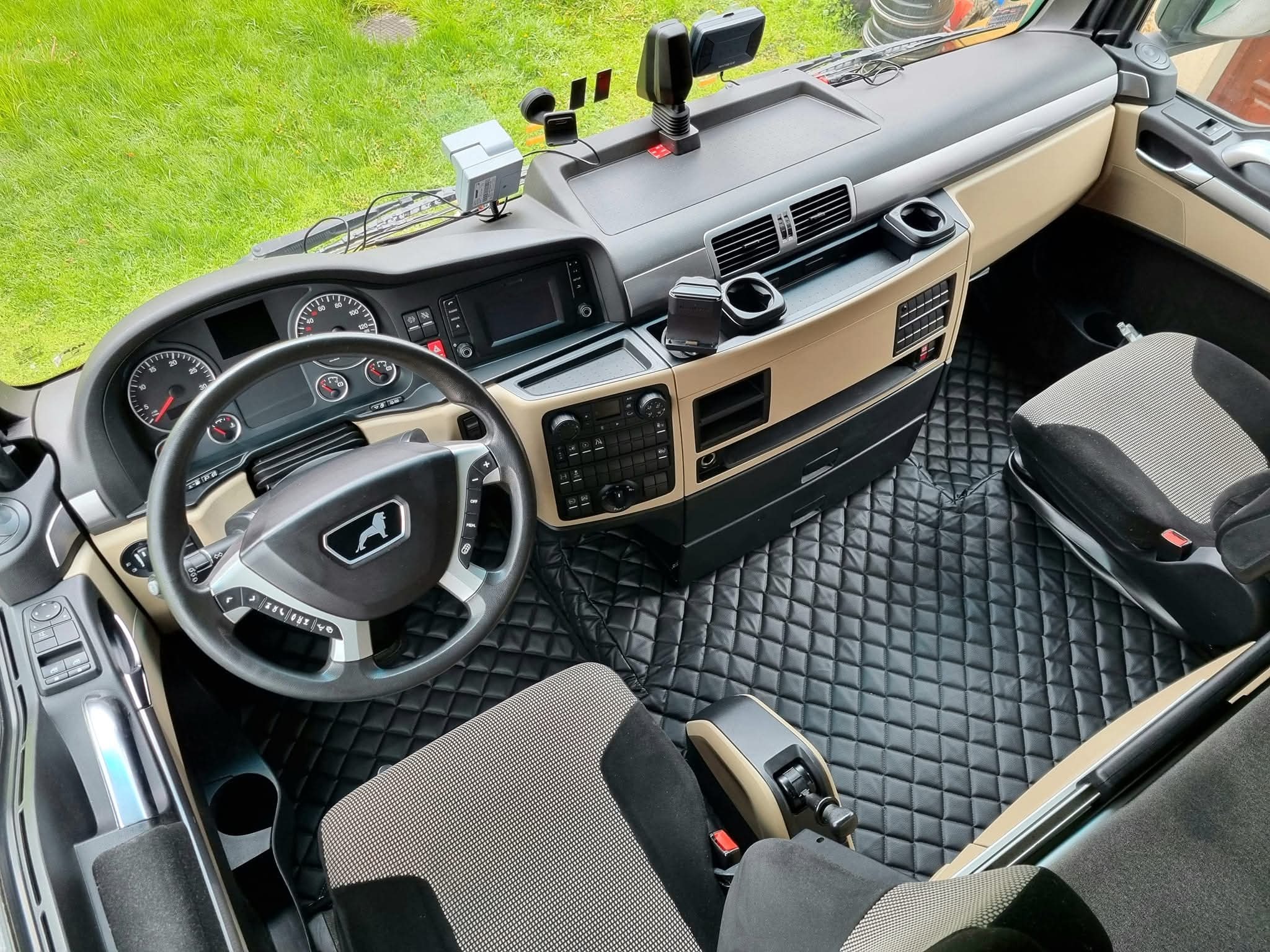 Wnętrze kabiny ciężarówki MAN z czarną, pikowaną skórą na podłodze i beżowymi elementami deski rozdzielczej. Widok z góry na kierownicę z logo lwa.