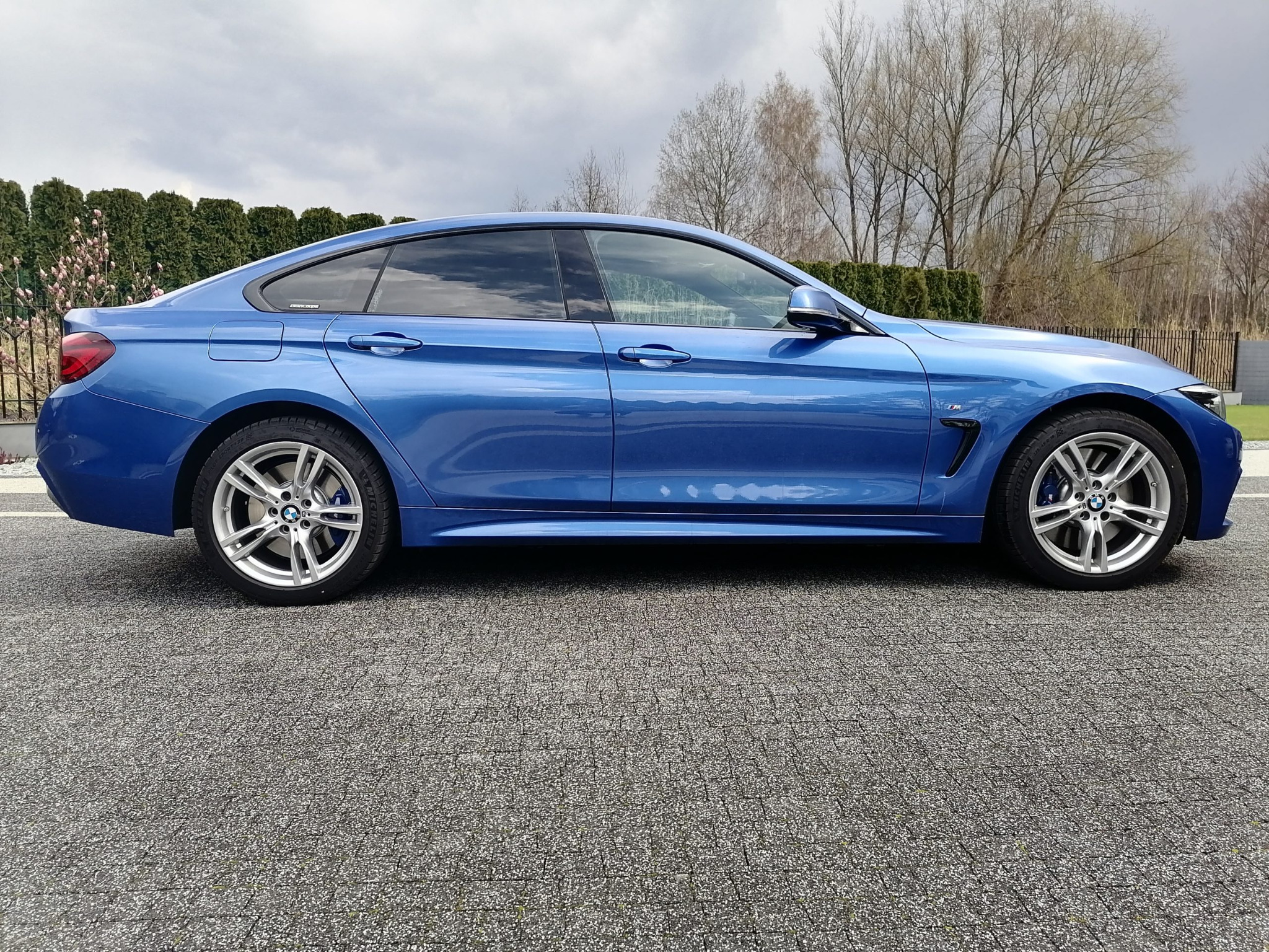 Błękitne BMW serii 4 Gran Coupe, widok z boku, z delikatnie przyciemnionymi szybami, zaparkowane na szarej kostce brukowej na tle żywopłotu i drzew bez liści.