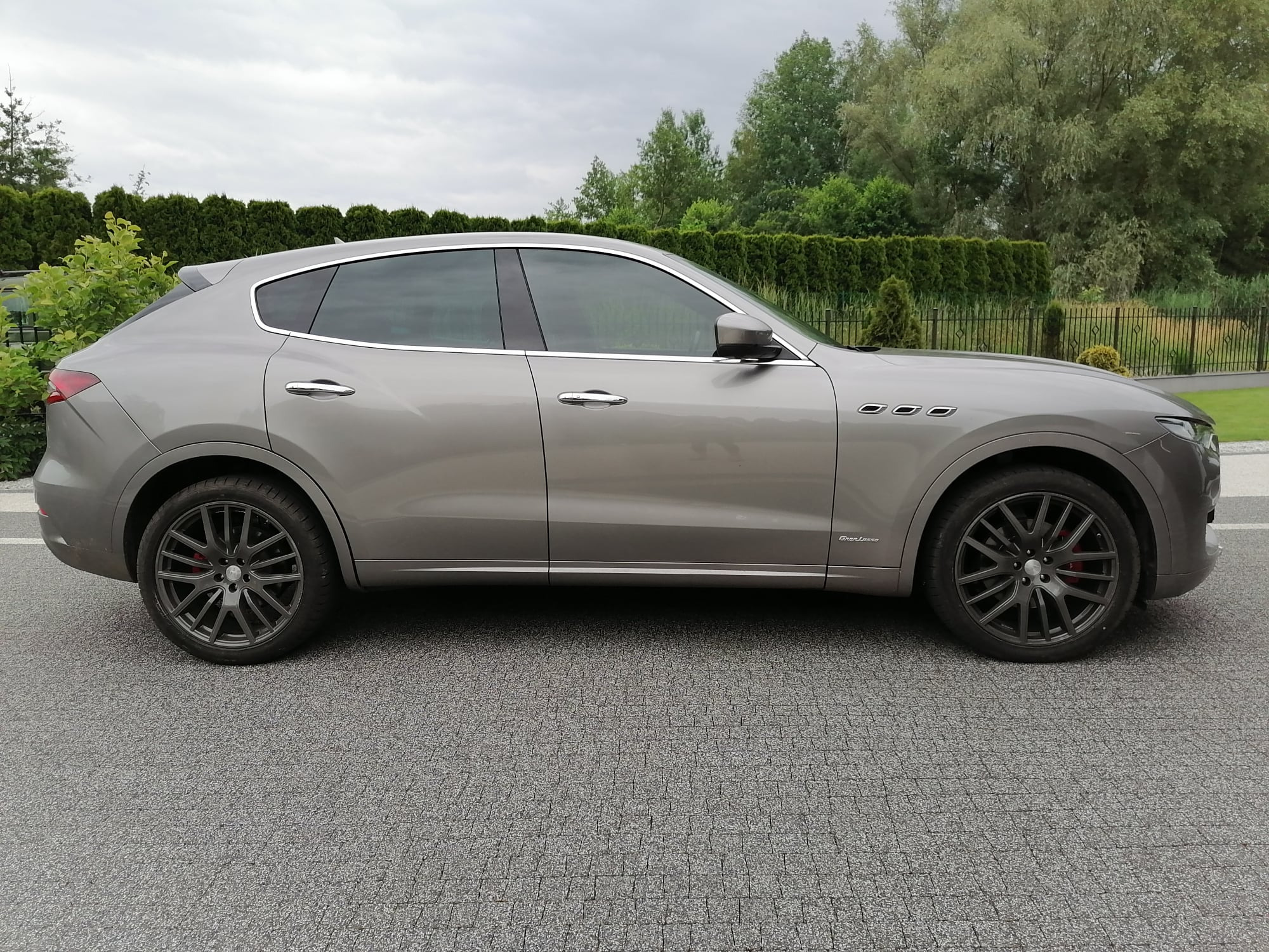 Szary samochód Maserati Levante na tle zielonego żywopłotu i pochmurnego nieba, widok z boku, felgi w kolorze grafitowym z czerwonymi zaciskami hamulcowymi.