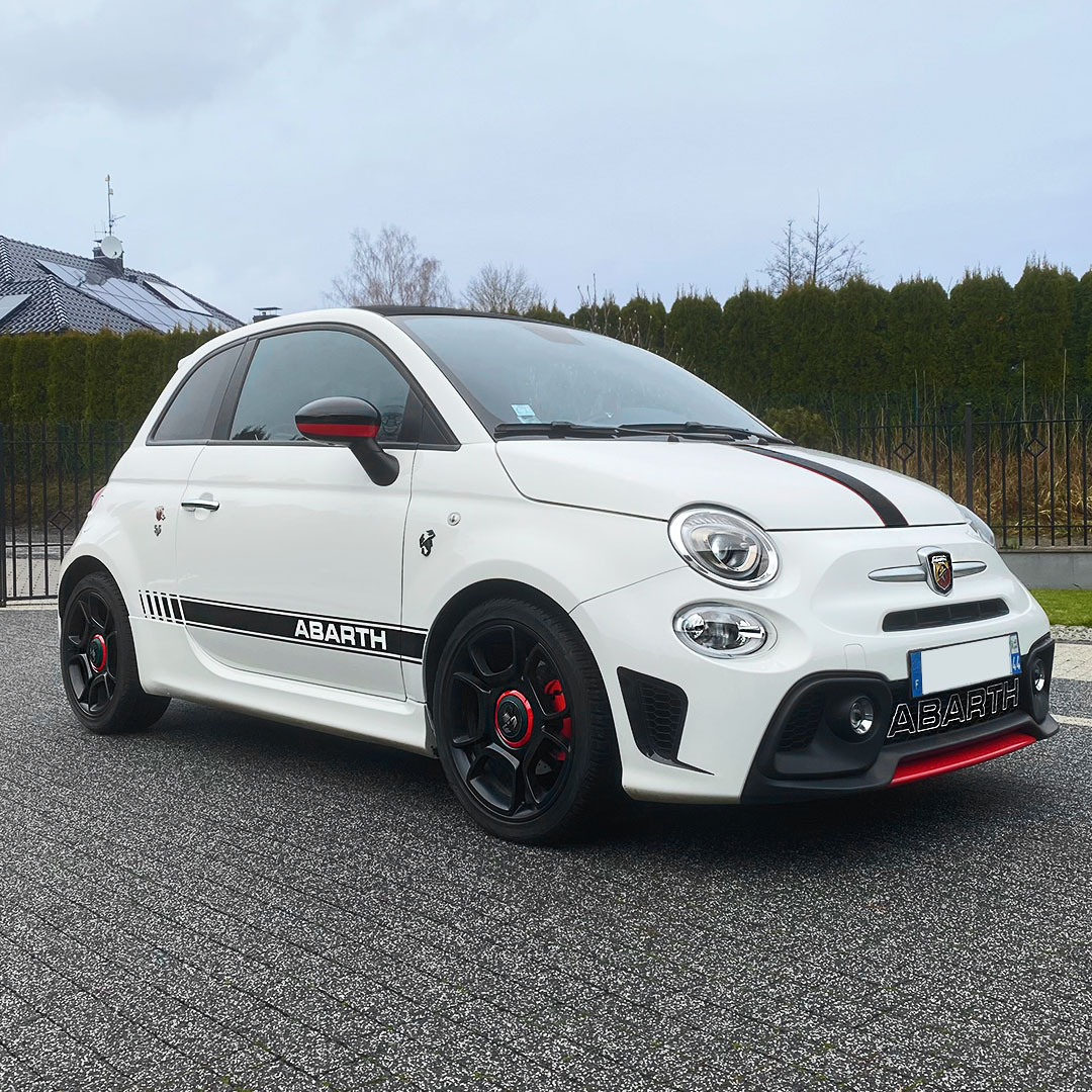 Biały samochód Abarth z czarnymi felgami z czerwonymi akcentami, czarnymi pasami na karoserii i masce, zaparkowany na kostce brukowej przed domem z zielonym żywopłotem.