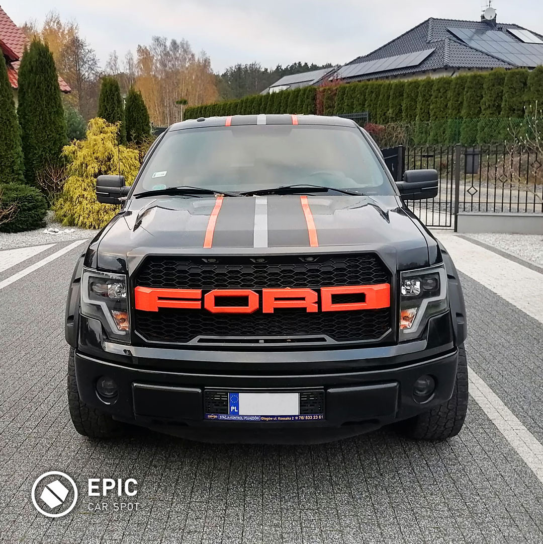 Czarny Ford Raptor z pomarańczowymi akcentami na grillu i pasami na masce, zaparkowany na szarym bruku przed domem z ciemnym dachem i zadbanym żywopłotem.