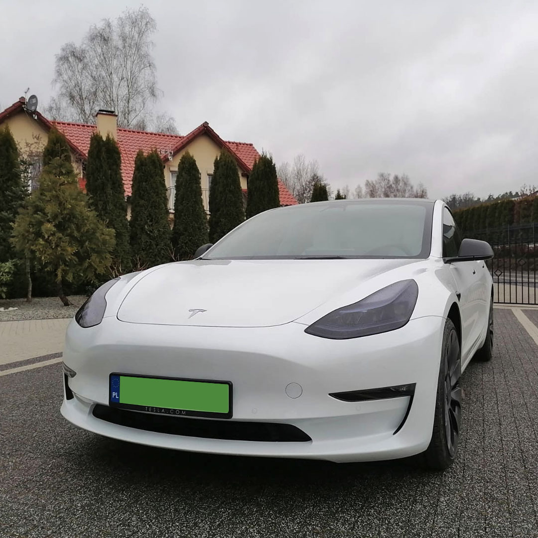 Biała Tesla Model 3 z przyciemnionymi szybami przednimi, zaparkowana na posesji z domem w tle.