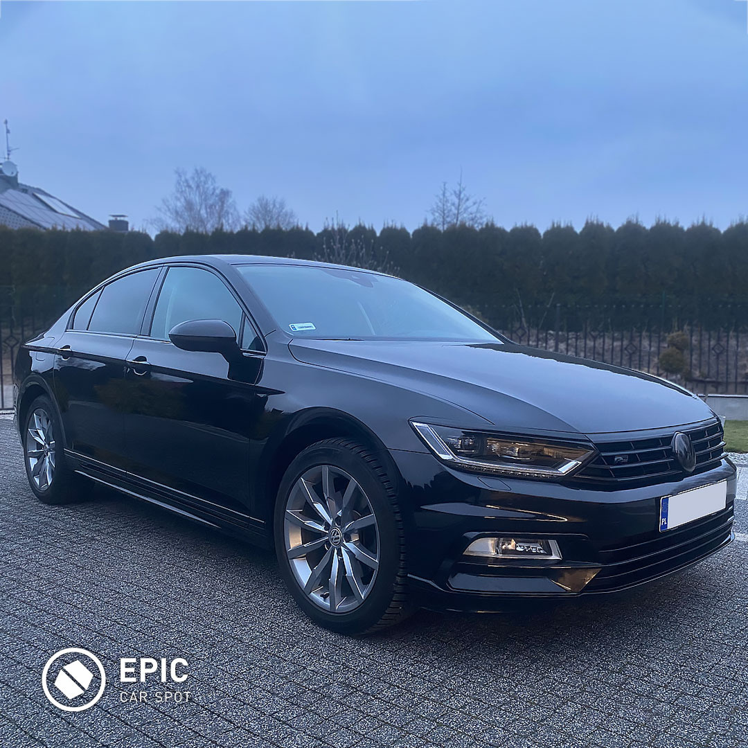 Czarny Volkswagen Passat z delikatnie przyciemnionymi szybami, zaparkowany na kostce brukowej. Widoczny fragment ogrodu w tle. Na przedniej szybie naklejka rejestracyjna.