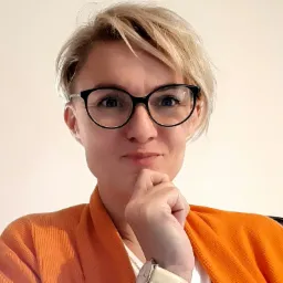 Portret kobiety w okularach i pomarańczowym kardiganie, z podpartą brodą, na jasnym tle.