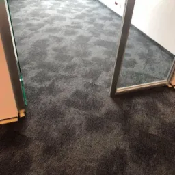 Szara, wzorzysta płytka dywanowa ułożona w biurze, widok z perspektywy wejścia, z widocznym grzejnikiem pod oknem w tle.