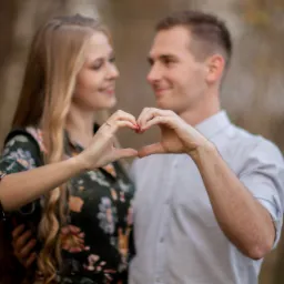 Zakochana para tworzy serce dłońmi na tle jesiennej scenerii, ona w kwiecistej sukience, on w jasnej koszuli.