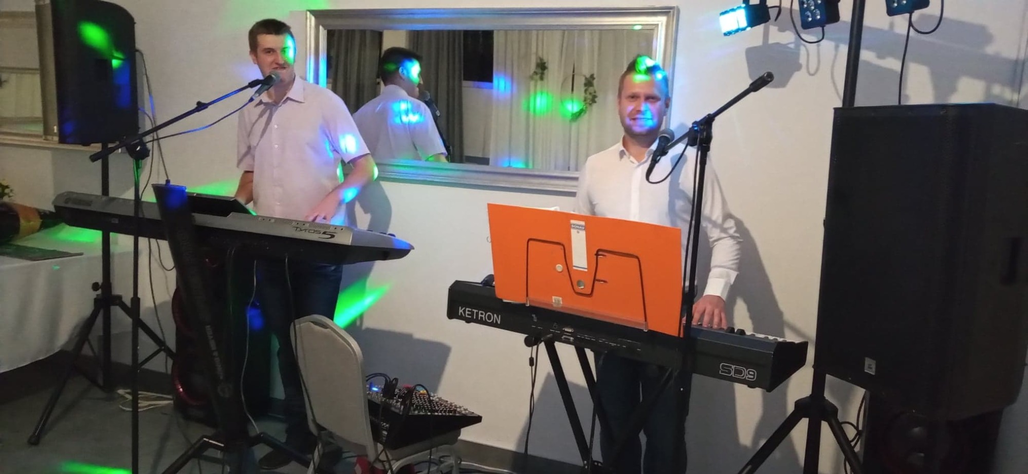 Dwóch mężczyzn gra na keyboardach Yamaha Tyros 5 i Ketron SD9, śpiewając do mikrofonów na tle białej ściany z lustrem i reflektorami.