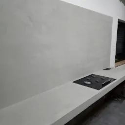 Szara ściana i blat kuchenny z wbudowaną płytą gazową, wykończone gładzią cementową, widoczne niedoskonałości na górze ściany.