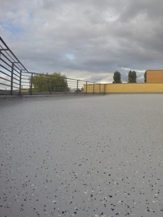 Szara, gładka powierzchnia z ciemnymi i jasnymi plamkami, zabezpieczona metalową balustradą, z widokiem na budynek z żółtej cegły i pochmurne niebo.