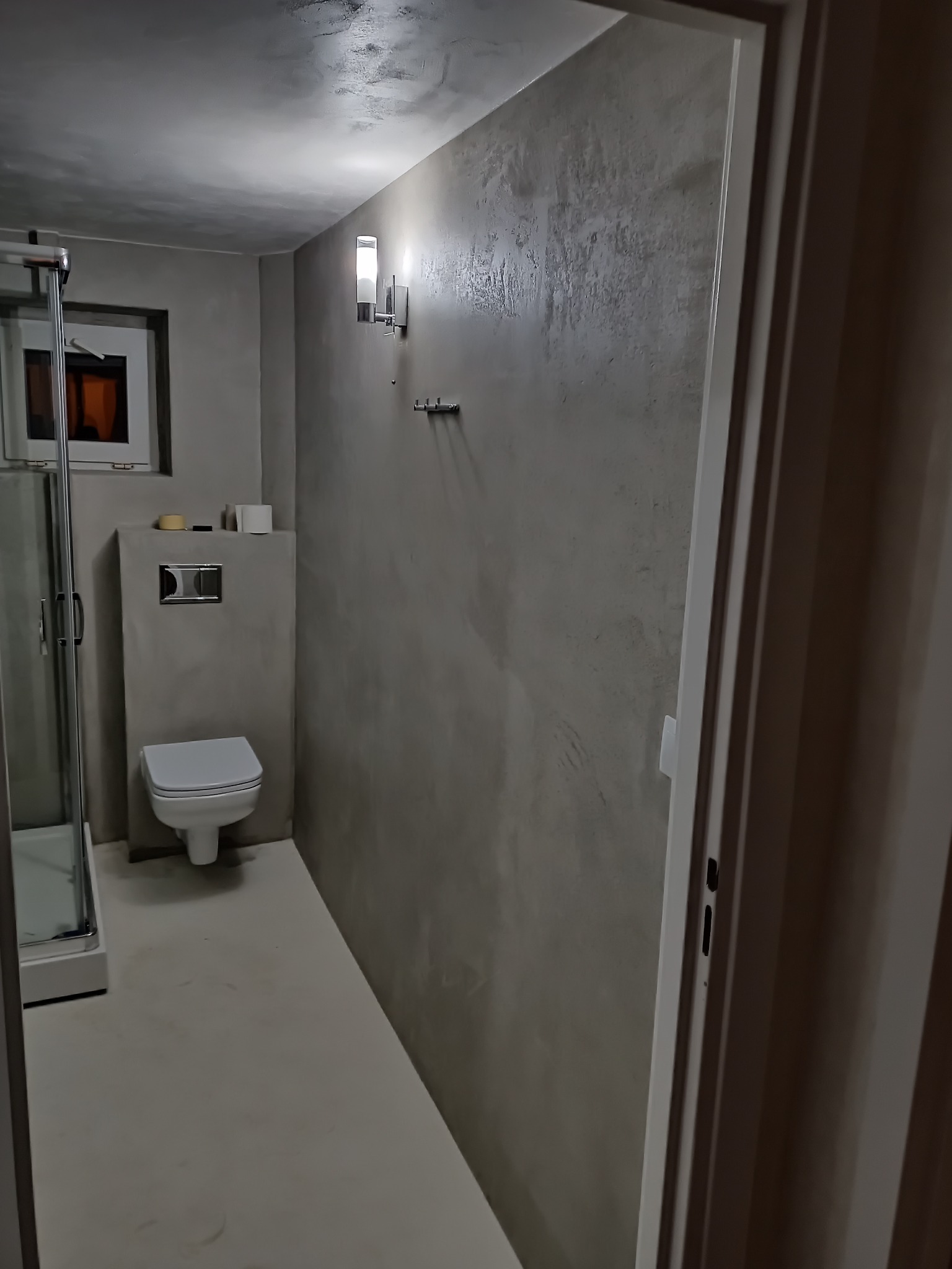 Nowoczesna łazienka w Bieruniu z betonową teksturą na ścianach, podświetlona lampami ściennymi, widok na kabinę prysznicową i toaletę.