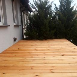 DREW-DOM Adrian Hauf - Nowo wybudowany drewniany taras przylegający do ściany budynku z widocznymi oknami i tujami w tle.