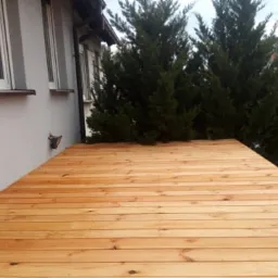 Nowo wybudowany drewniany taras przylegający do ściany budynku z widocznymi oknami i tujami w tle.