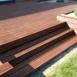 DREW-DOM Adrian Hauf - Drewniany taras z trzema stopniami schodów, zakończony donicą z roślinami, oświetlony słonecznym światłem.