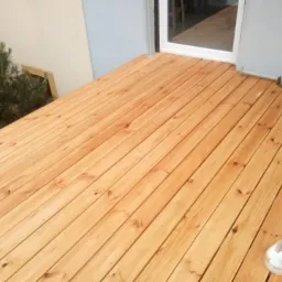 Świeżo położony drewniany taras z jasnego drewna, widok z góry, przy ścianie budynku z oknem, obok leży pojemnik z wkrętami.