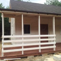 DREW-DOM Adrian Hauf - Surowy drewniany dom z gontowym dachem i zadaszonym gankiem z białą balustradą, w otoczeniu drzew iglastych.