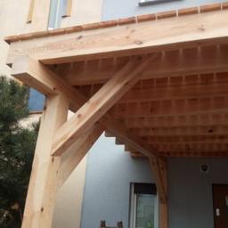 DREW-DOM Adrian Hauf - Drewniana konstrukcja z belek tworząca zadaszenie nad wejściem do domu, widoczne elementy dachu i wspierające słupy.