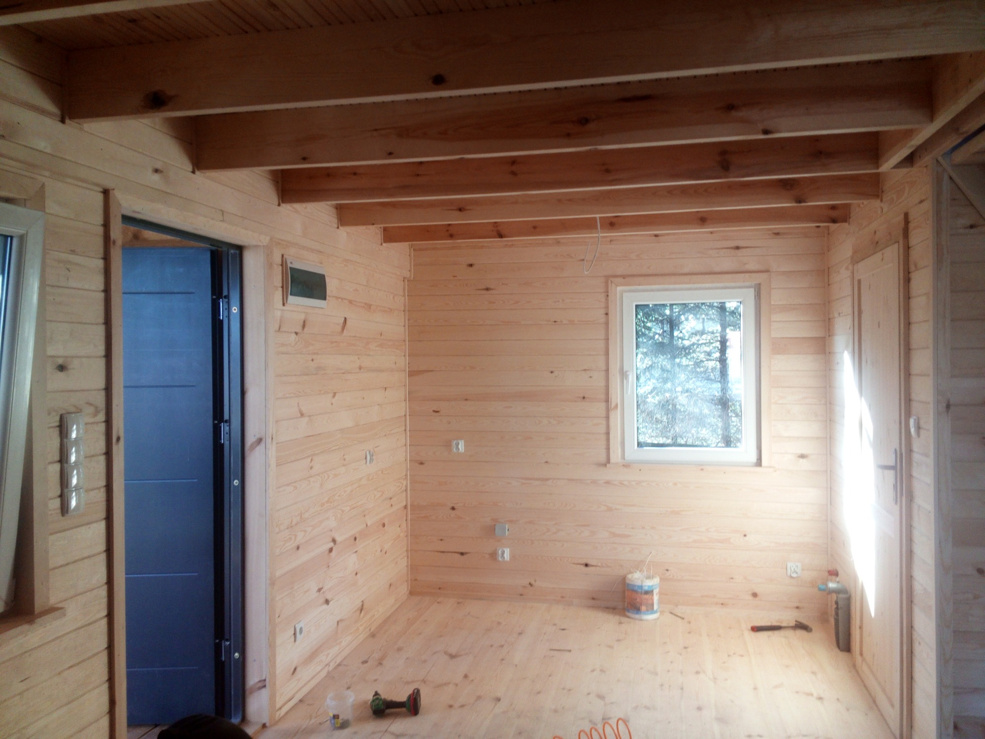 Wnętrze surowego drewnianego domu z widocznymi belkami stropowymi, niebieskimi drzwiami i narzędziami na podłodze, w trakcie wykańczania.