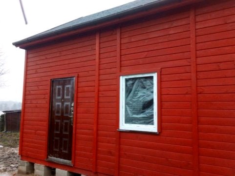 Czerwony dom szkieletowy w Poznaniu, z widocznym wejściem i oknem zabezpieczonym folią, posadowiony na betonowych bloczkach.