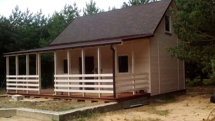 Niewielki, parterowy dom z drewna z zadaszonym tarasem, brązowym dachem dwuspadowym i białymi balustradami, otoczony drzewami.