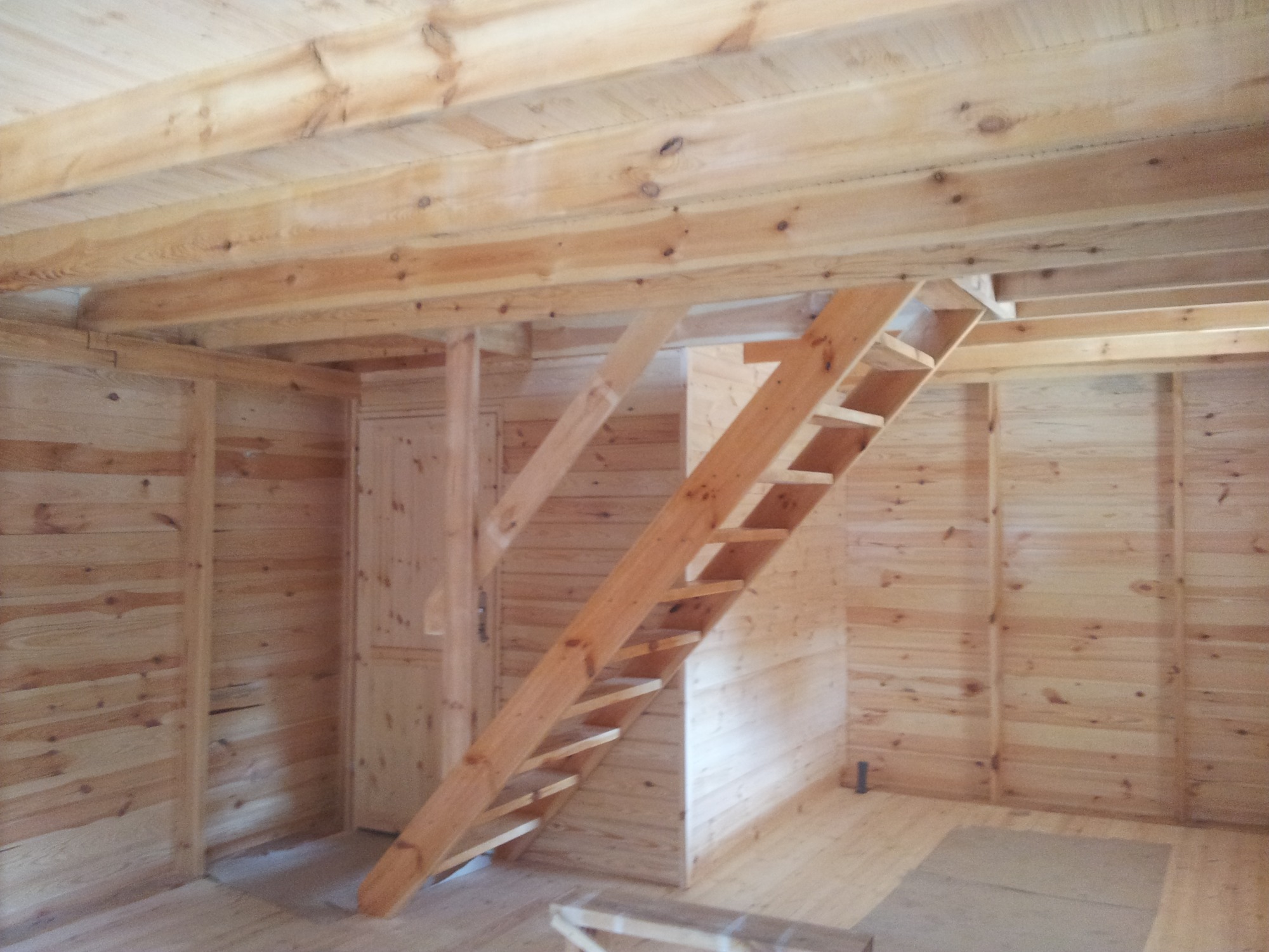 Wnętrze surowego drewnianego domu w trakcie budowy, widoczne drewniane ściany, strop i schody drabiniaste.
