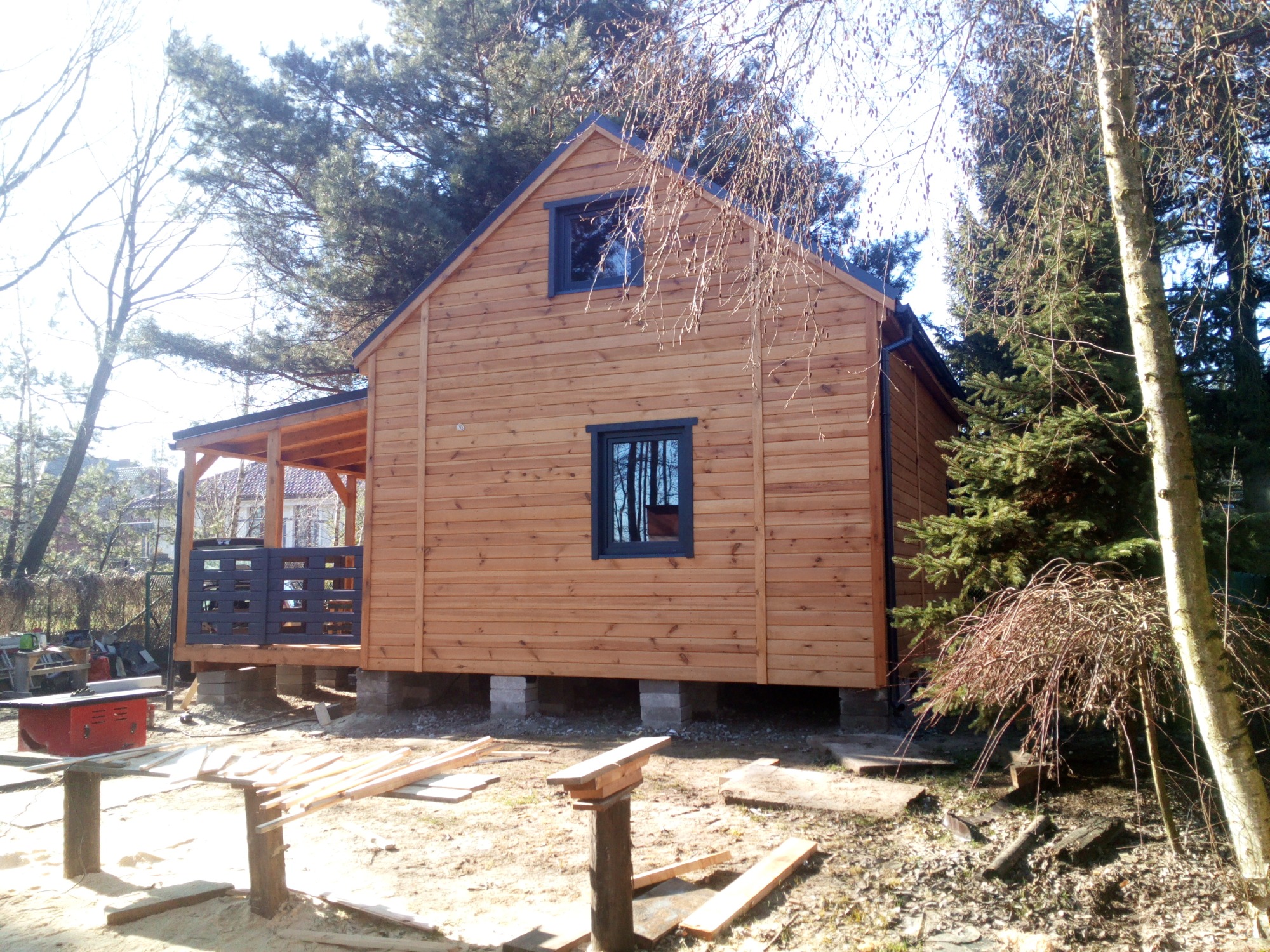Surowy, drewniany domek letniskowy z zadaszonym tarasem, posadowiony na betonowych bloczkach, w otoczeniu drzew i z widocznymi narzędziami budowlanymi na pierwszym planie.