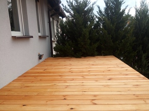 Nowo wybudowany drewniany taras przylegający do ściany budynku z widocznymi oknami i tujami w tle.