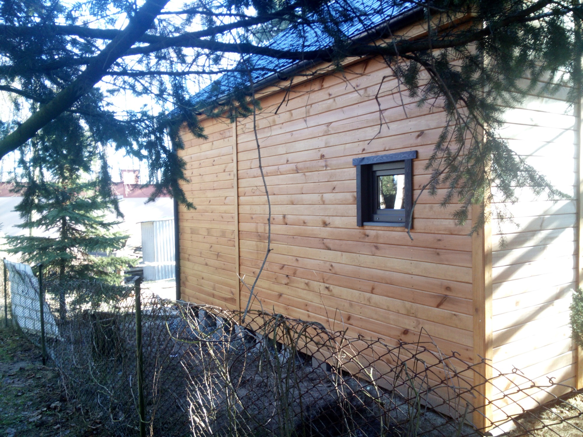 Ściana zewnętrzna domu z drewnianych paneli, widoczna przez gałęzie drzewa i metalową siatkę ogrodzeniową.