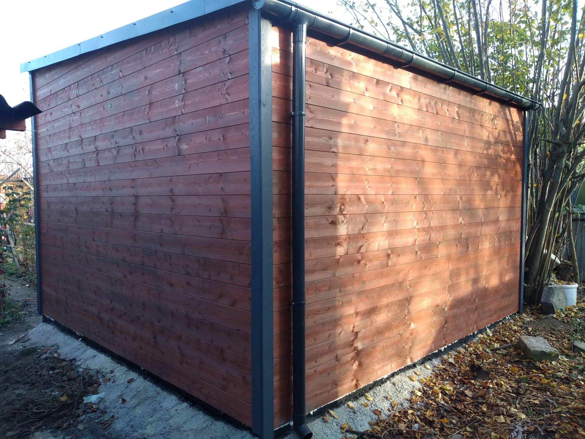 Gotowa drewniana konstrukcja szkieletowa z ciemnobrązowymi deskami, widoczne rynny i obróbki blacharskie, teren wokół wysypany żwirem i liśćmi.
