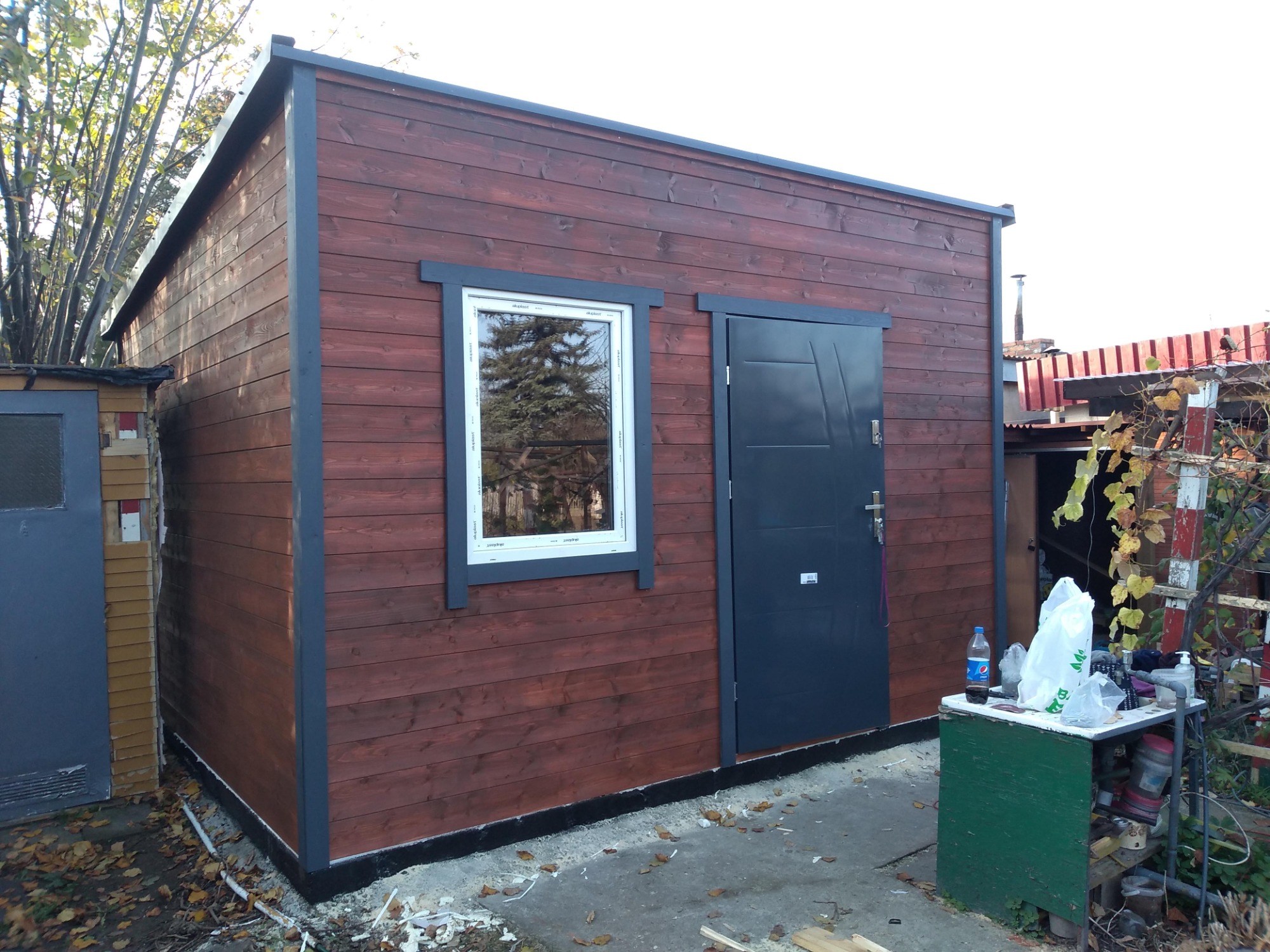 Gotowy, drewniany budynek o konstrukcji szkieletowej z szarymi obramowaniami okna i drzwi, widoczny w ogrodzie.