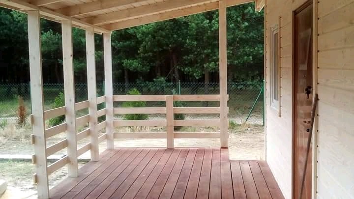 Zadaszony drewniany taras przy domu z bali, widok z zewnątrz na las w tle, widoczne elementy konstrukcyjne i drewniana podłoga tarasu.